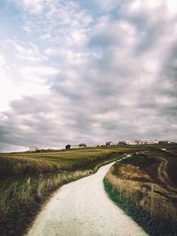 Dirt road passing through field against cloudy sky