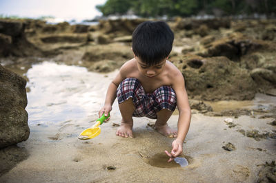 Shirtless boy playing with sand while crouching at beach