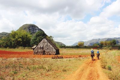 Rear view of men riding horses by hut against cloudy sky