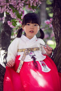 Korean girl child in a national costume sit on tree branch in a garden with cherry blossoms 