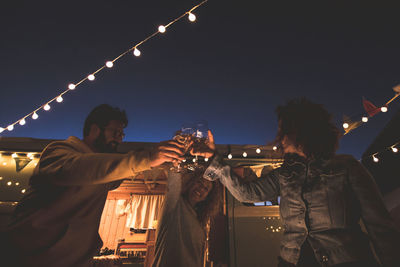 Male and female friends toasting while standing against sky at night