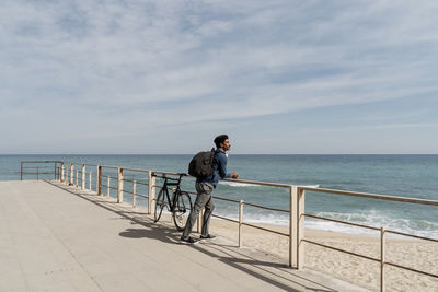 Mid adult man carrying backpack while leaning on railing by bicycle at beach during sunny day