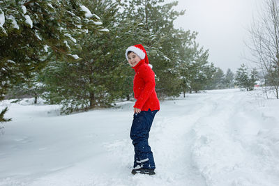 A happy teenager in a red sweater  walks in the winter in the park, near the pines in the snow