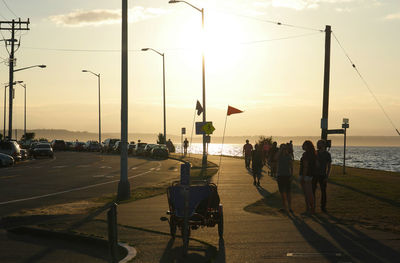 People walking on sidewalk by alki beach against sky during sunset