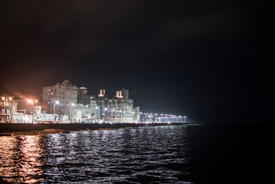 Illuminated buildings by sea against sky at night