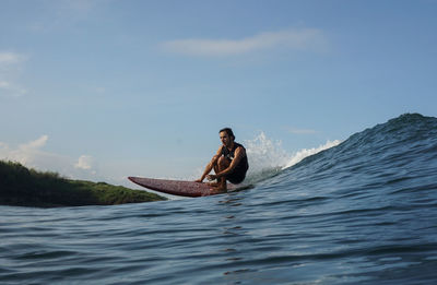 Man sitting on surfboard in sea against sky