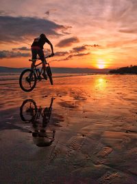 Silhouette man riding bicycle on beach against sky during sunset