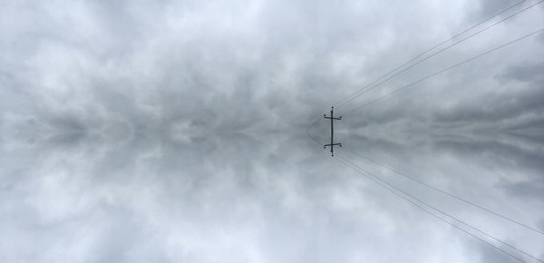 Reflection of electricity pylon against cloudy sky on calm lake