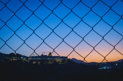 Silhouette cityscape behind chain link fence against sky during sunset