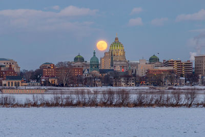 Illuminated buildings in city against moon during winter