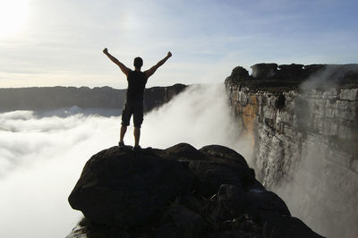 Rear view of man with arms raised standing on rock against sky