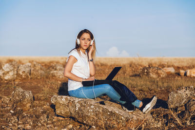 Young woman with laptop sitting on land against sky