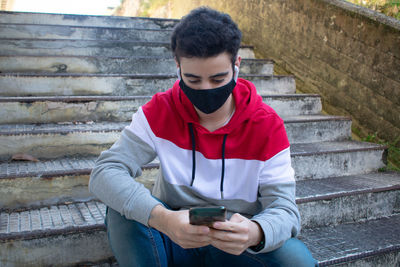 Young caucasian man wearing a black face mask and casual clothing, sitting on some stairs, with earb