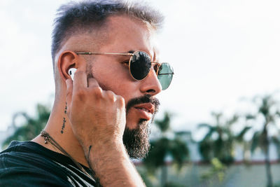 Side view of thoughtful bearded male with tattoos in casual clothes and sunglasses wearing earphones and looking away on street against green trees in sunny day