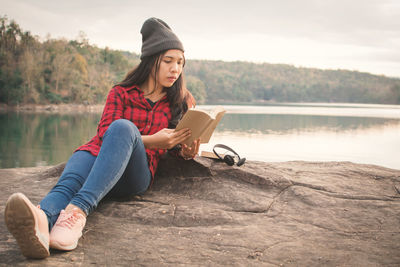 Young woman lying on rock reading book by lake against clear sky
