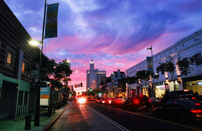 Traffic of city street by buildings against sky during dusk