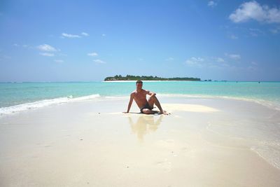 Full length portrait of shirtless man standing at beach against blue sky