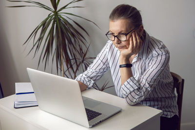 Woman in a white striped shirt aged sits at a computer at home with glasses