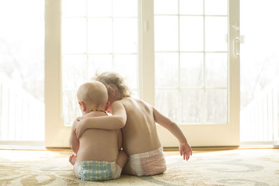 Rear view of shirtless siblings sitting by window at home