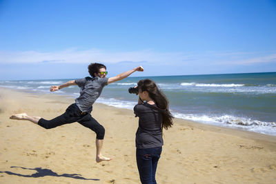 Woman photographing man jumping at beach against sky