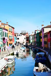Boats moored on canal amidst buildings against blue sky