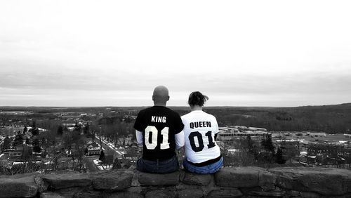 Rear view of couple sitting on retaining wall against sky