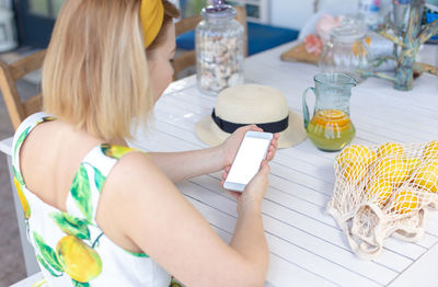 A blonde girl sits at a white table, holds a white smartphone, next to it is a jug of lemonade 