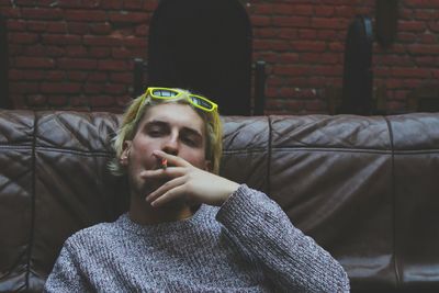 Portrait of man smoking cigarette while sitting on sofa against brick wall