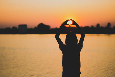 Silhouette woman making heart shape with hands by lake against sky during sunset