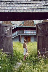 Blonde village girl with long hair stands in a wooden gate in the village in summer