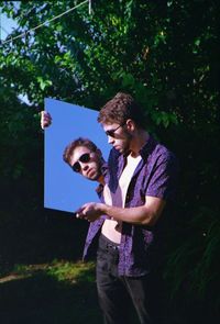 Young man holding mirror while standing against tree