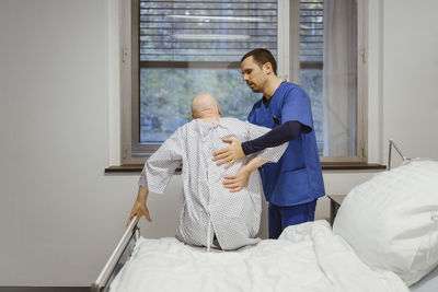 Male nurse helping senior patient sitting on bed at hospital