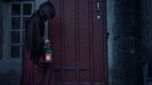 Side view of woman standing against closed door holding a light lantern