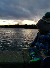 Man sitting by lake against sky during sunset