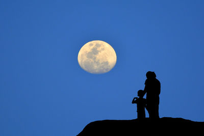 Low angle view  silhouette of a mother and son watching the moon on mountain orange background