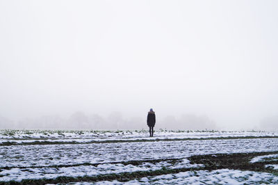 Mid distant rear view of person standing on snow covered field against sky during winter