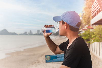 Man drinking water from a bottle sit outside next to the beach.