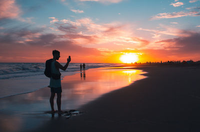 Silhouette man standing on shore at beach against dramatic sky during sunset