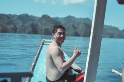 Portrait of shirtless cheerful young man gesturing thumbs up on boat in sea