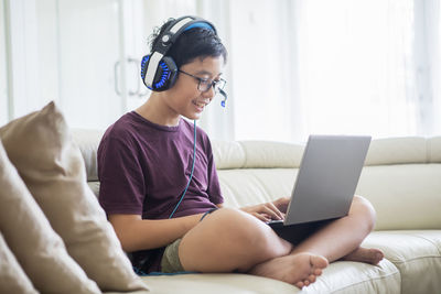 Smiling boy wearing headphones while using laptop on sofa