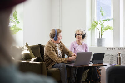 Senior man and woman talking to each other while sitting in living room and using laptop and digital tablet to edit podcast