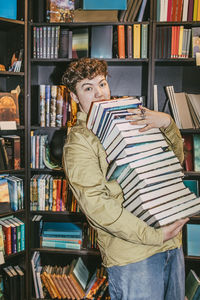 Portrait of young man balancing heap of books near shelf in bookstore