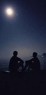 Silhouette people sitting on shore against sky at night