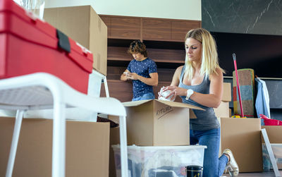 Woman unpacking cardboard box while man using mobile phone while standing at home