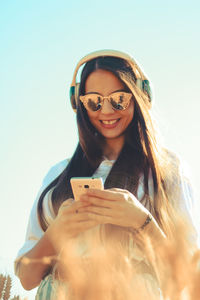 Woman listening music while using mobile phone against clear sky