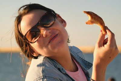 Close-up of young woman with sunglasses eating food against sky during sunset