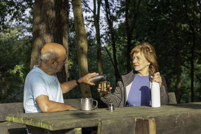 Senior man pouring water from a reusable bottle into a metallic mug to his wife while sitting