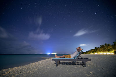 Man lying on sunbed on sand beach and looking up to stars on night sky.