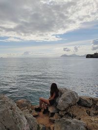 Full length of young woman sitting by rock against sea