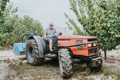 Side view of senior male farmer driving tractor amidst fruit trees during work in orchard on summer day